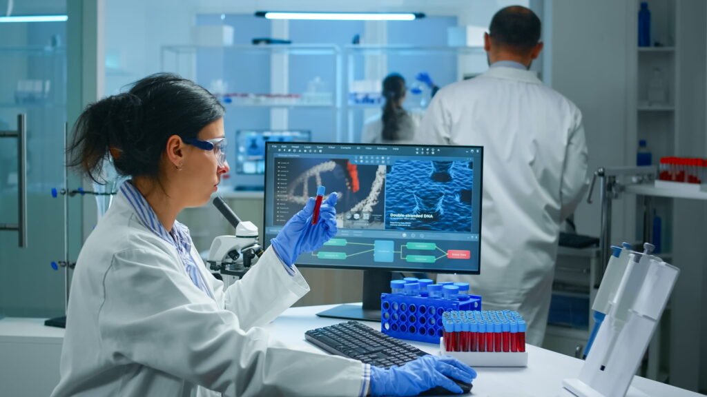 A researcher in a lab reviewing DNA on a monitor while handling sample tubes at her workstation.